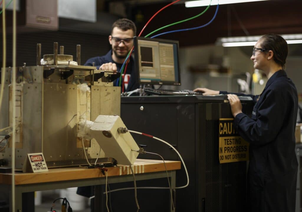 Two engineers collaborating at a laboratory testing station, monitoring real-time data from a high-temperature furnace during advanced material performance characterization.