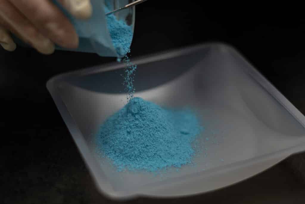 Researcher pouring blue precursor powder for advanced material synthesis and formulation.