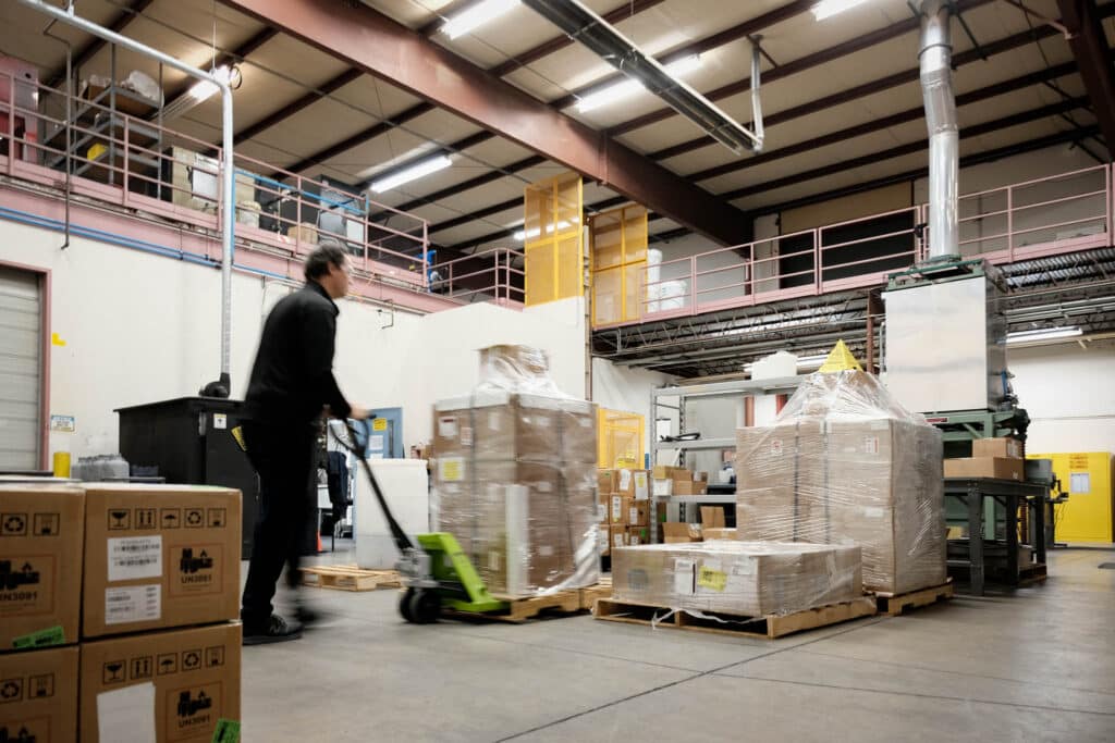 A technician using a pallet jack to move shrink-wrapped pallets of finished products in a large industrial warehouse, demonstrating commercial-scale manufacturing and logistics capabilities.