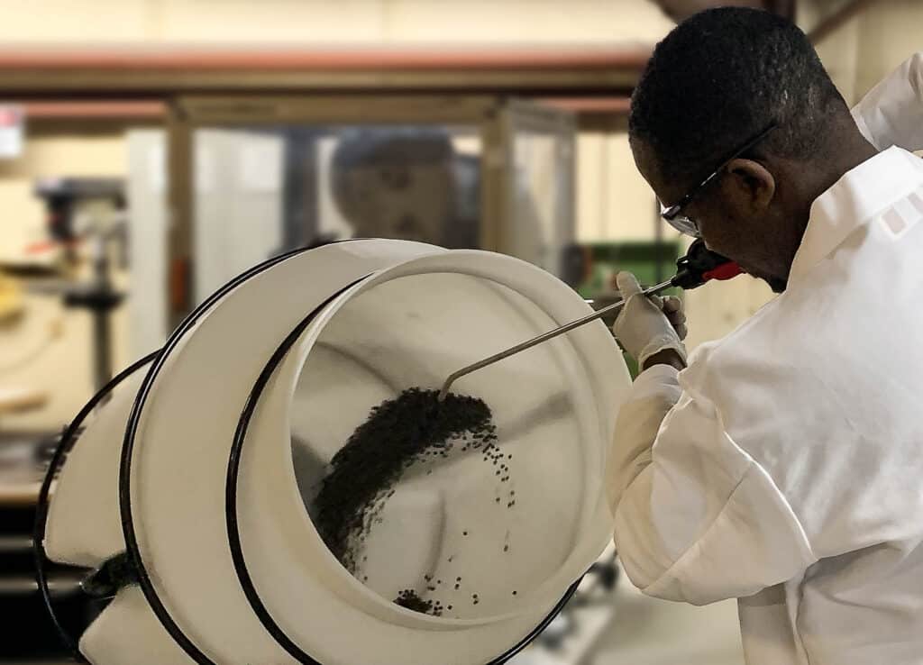 Technician using a rotary drum to apply a specialized coating to advanced catalyst pellets during the material scale-up process.